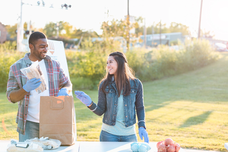 A young woman smiles as her friend while volunteering together at a community food drive. They are filling a paper bag with food donations.